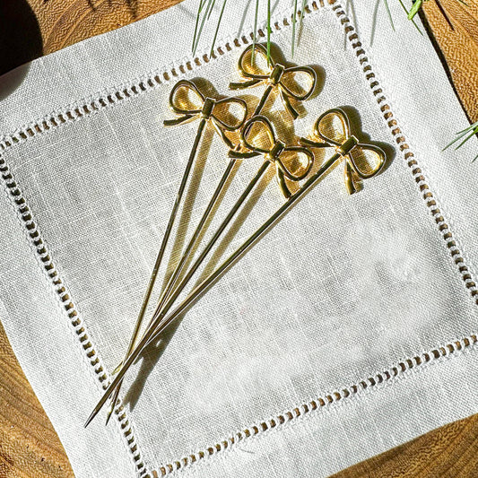 Four gold-plated, bow-shaped cocktail picks from the Threads & Honey LIMITED RELEASE are elegantly arranged on a white linen napkin with decorative stitching. These entertaining accessories fan out gracefully on a textured wooden surface, with sunlight casting soft shadows.